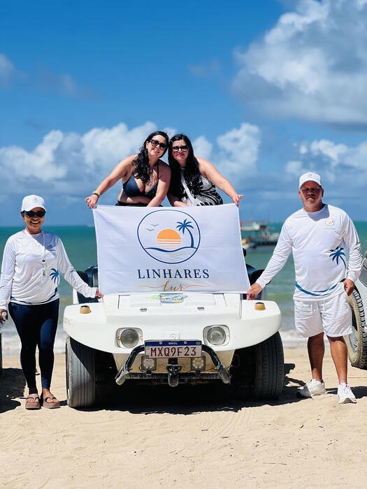 Grupo segurando a bandeira da Linhares Tur ao lado de um buggy branco na praia